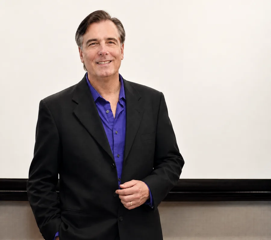 Toby Martini smiling in a black jacket and purple shirt against a warm white background.