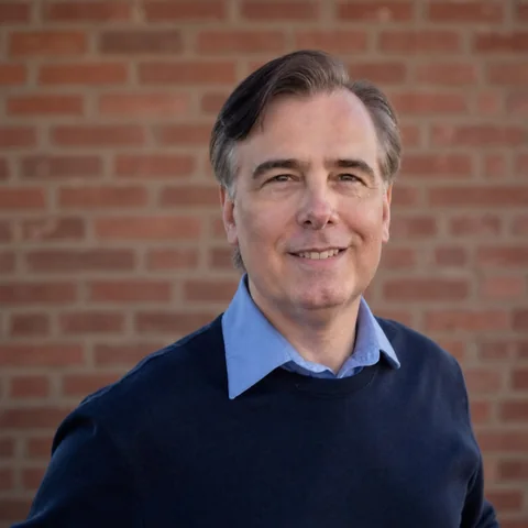 Toby Martini smiling in a blue sweater in front of a brick wall.